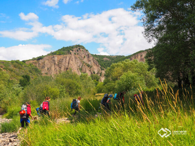 Uferlandschaft an der Nahe, den Rotenfels im Blick, im Vordergrund wandern mehrere Packrafterinnen mit Rucksäcken zum Einstieg in den Packrafting Erlebnistag Nahe