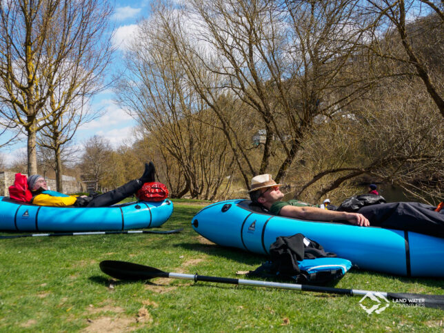 Zwei Personen liegen in zwei hellblauen Packrafts auf einer Wiese in der Sonne