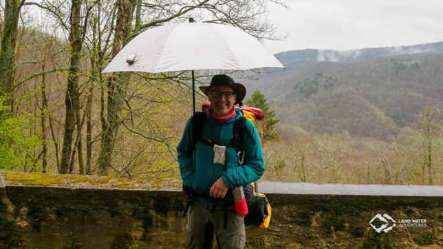 Eine Person steht mit Regenschirm und großem Rucksack bepackt vor einer Mauer, im Hintergrund das Hahnenbachtal