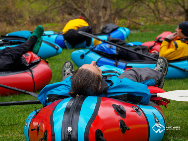 Eine Person liegt auf einem Packraft auf einer Wiese und ruht sich aus. Im Hintergrund weitere Boote.