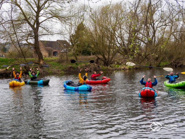 Sechs Personen in bunten Packrafts auf dem Fluss werfen freudig strahlend ihre Paddel in die Luft.