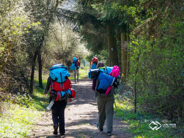 Mehrere Personen mit großen, bepackten Rucksäcken wandern einen schmalen Weg entlang, es ist Frühling.