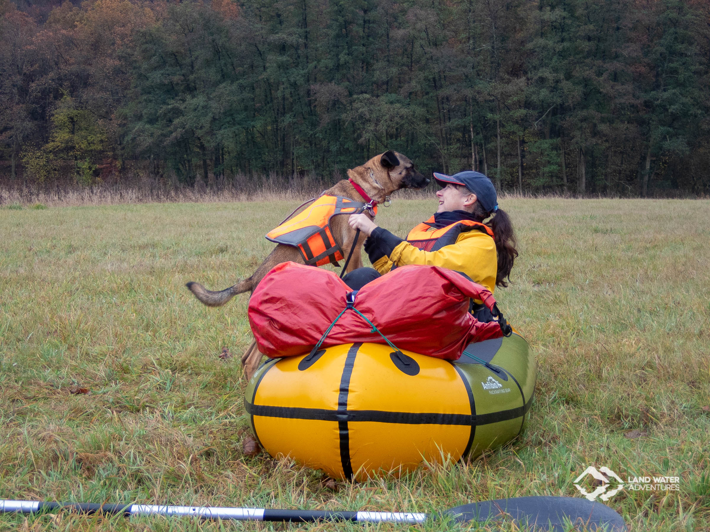 Eine Frau sitzt auf der Wiese im bunten Packraft, während der Hund zu ihr ins Boot klettern möchte